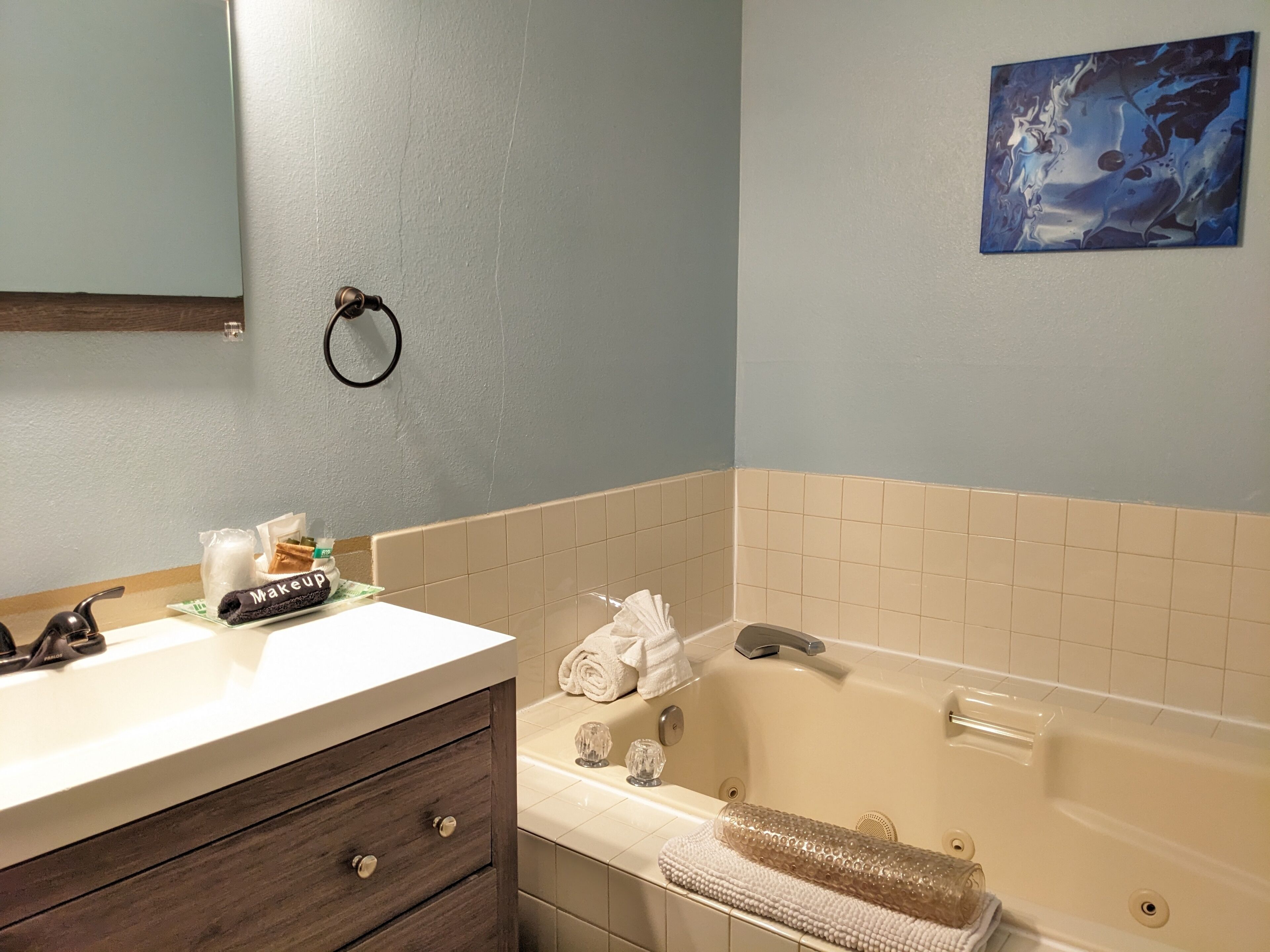 Un baño con un lavamanos blanco, tocador de madera oscura, bañera color beige y un cuadro abstracto azul en la pared.