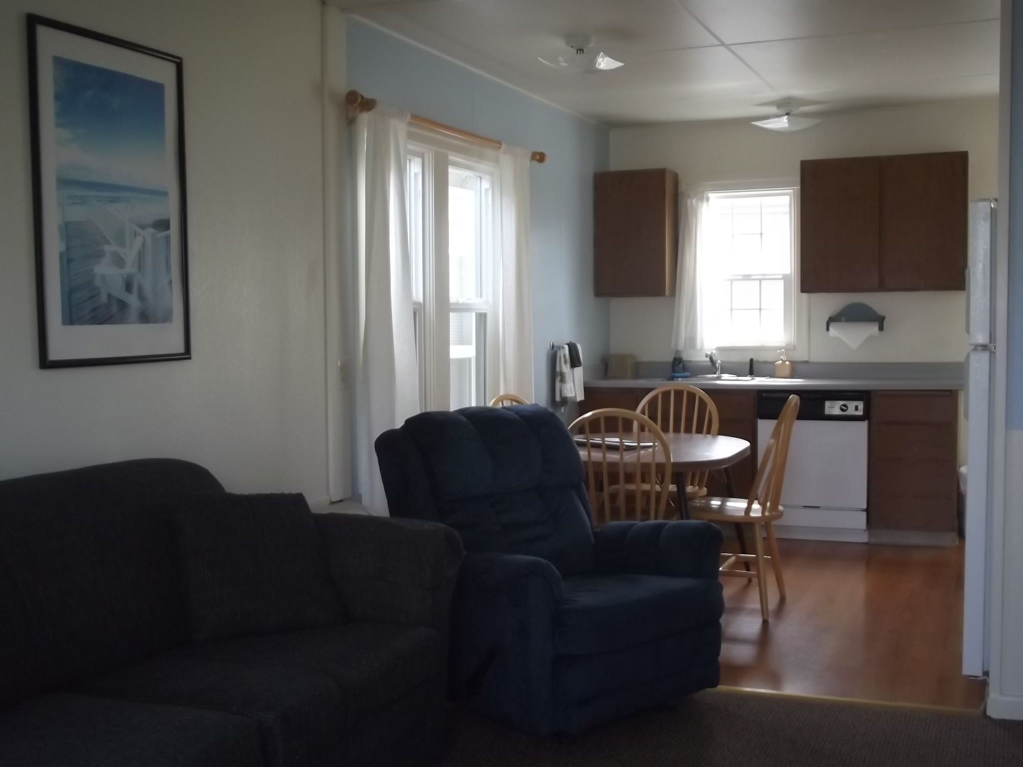 A living room with a dark sofa and a blue armchair, a dining area with wooden chairs and a table, and a kitchen with wooden cabinets and a dishwasher.