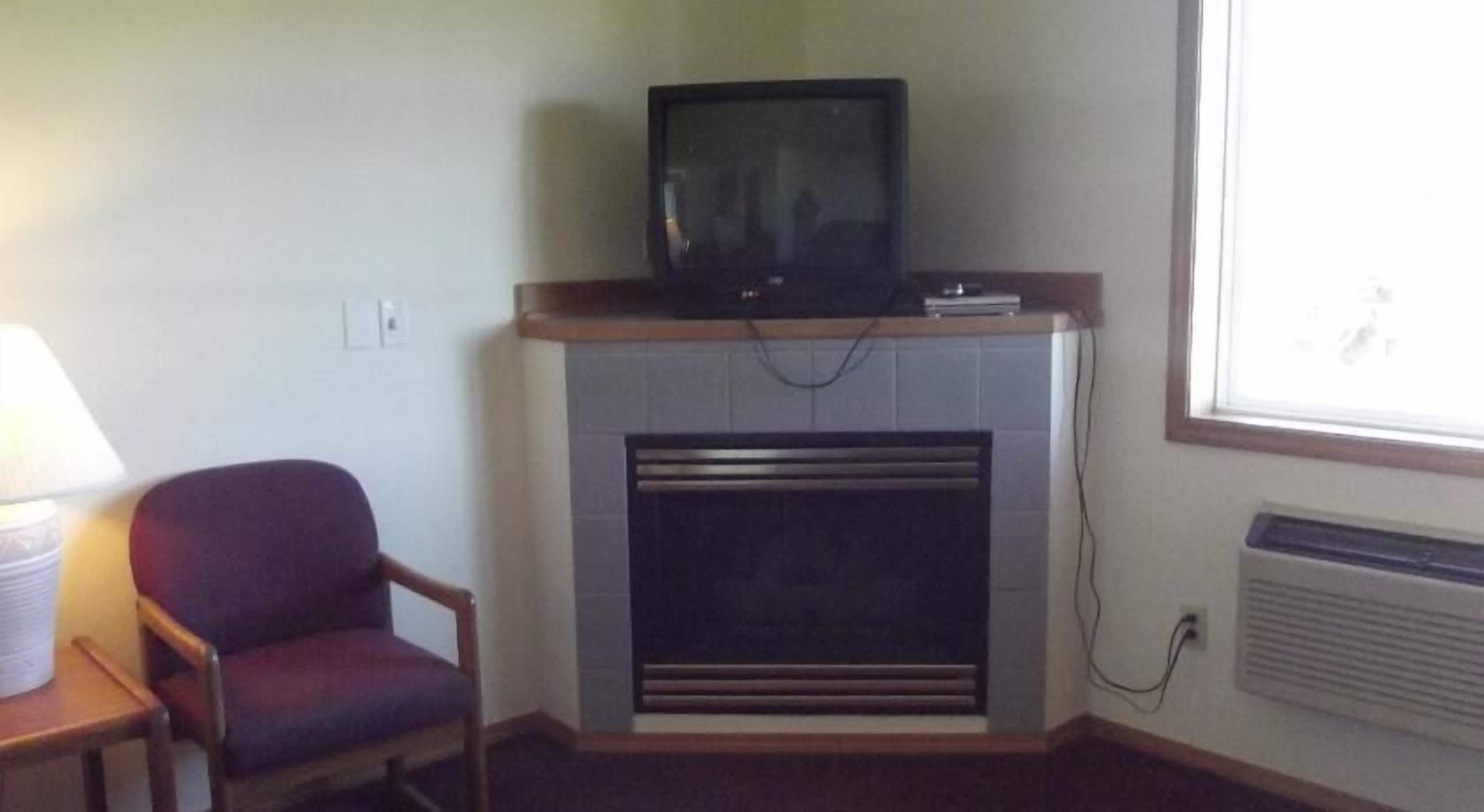 A room with a television on a stand, a chair, a fireplace, and a window.