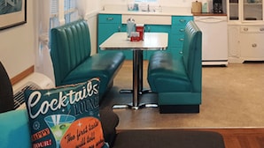 Una cocina con armarios color turquesa, una encimera blanca y una mesa de comedor con sillas color turquesa. Hay una lámpara colgante sobre la mesa.