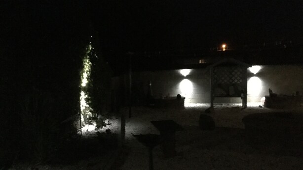 Un espacio exterior tenuemente iluminado con un pequeño árbol iluminado, una zona cubierta con asientos y algunas mesas y sillas.
