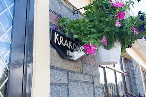 Un bâtiment sur lequel on peut lire l’inscription « Krakow 190 », et un pot de fleurs suspendu contenant des fleurs roses et violettes.