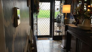 Un pasillo con baldosas de diseño, una puerta negra que da al exterior, un taburete pequeño y una mesa con una lámpara y un jarrón de flores.