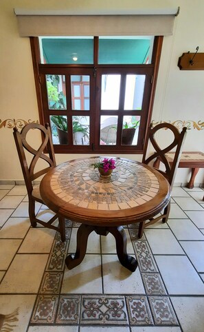 Une table ronde dont le plateau est recouvert d’une mosaïque, entourée de quatre chaises en bois aux dossiers finement sculptés. Devant la table, on aperçoit une petite plante en pot ornée de fleurs violettes.