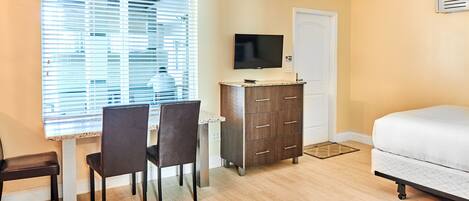 A bedroom with a bed, a TV on a dresser, a desk with chairs, and a window with blinds.