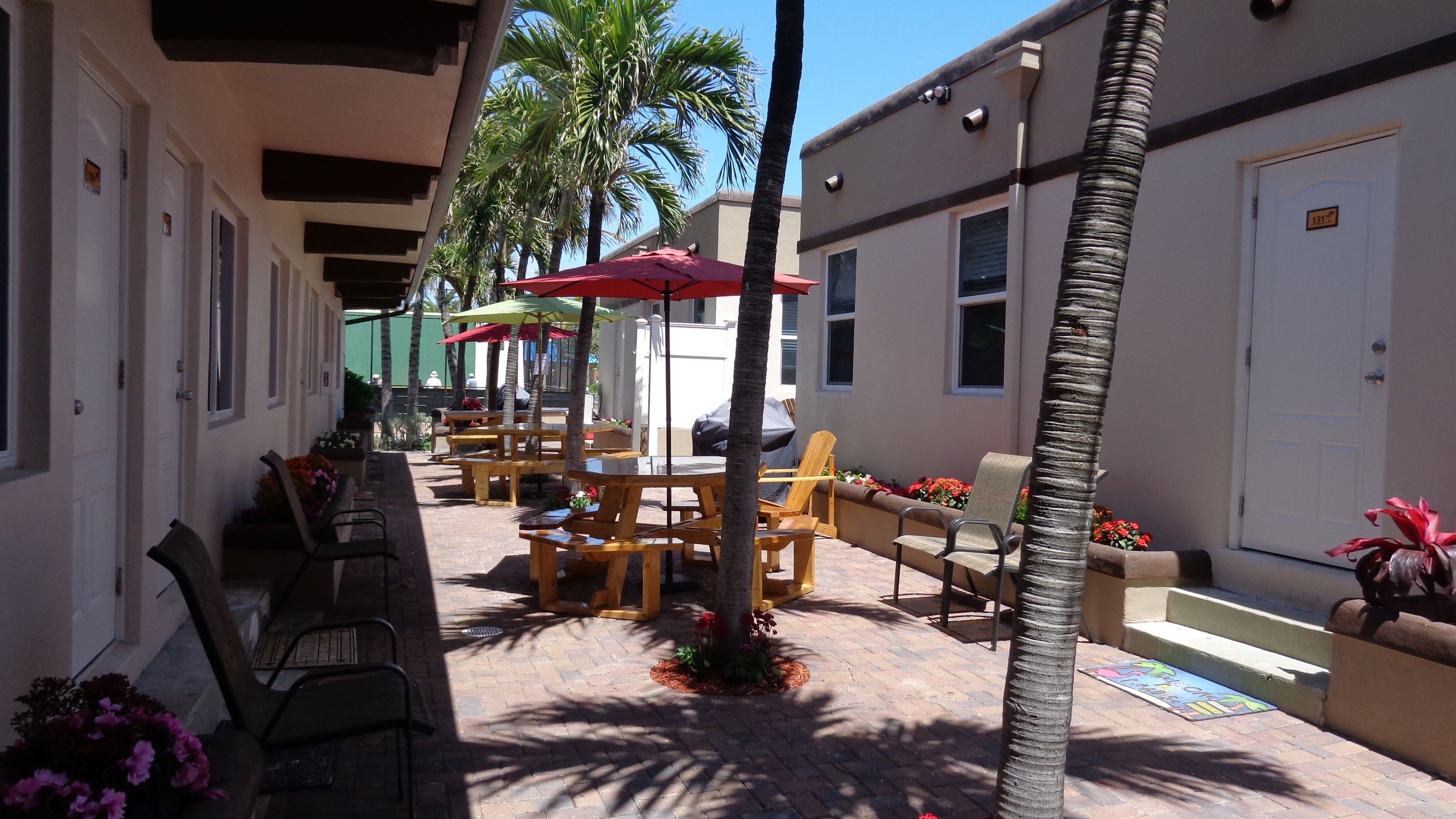 An outdoor patio area with wooden tables and chairs, palm trees, and a covered seating area with red umbrellas.