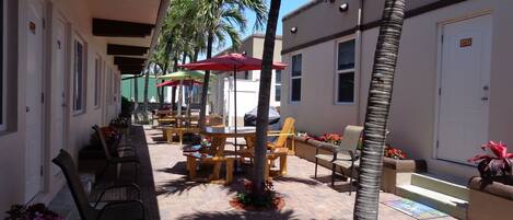 An outdoor patio area with wooden tables and chairs, palm trees, and a covered seating area with red umbrellas.