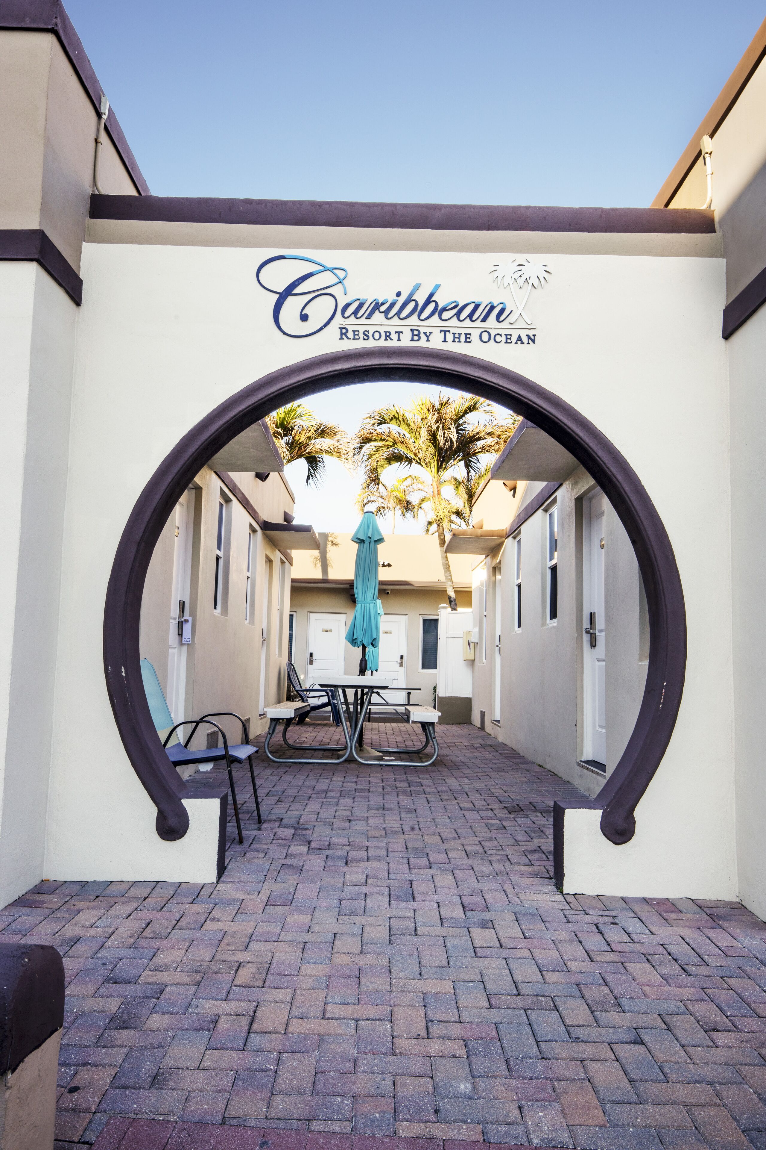 Un arco conduce a un cortile con un tavolo e sedie, un ombrellone color turchese e un cartello con la scritta "Caribbean Resort By The Ocean".