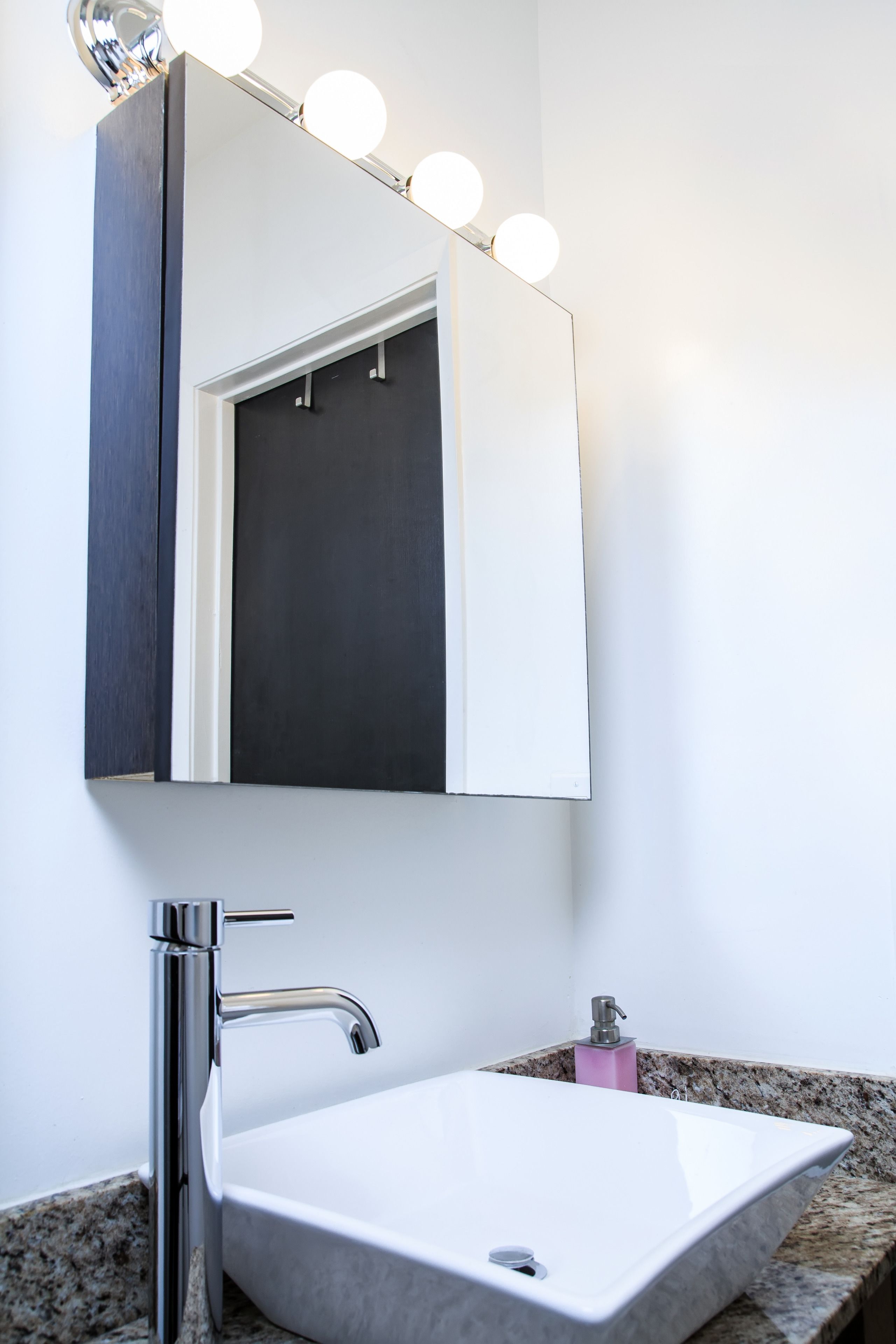 A bathroom with a white sink, a chrome faucet, a mirror with lights, and a dark cabinet.