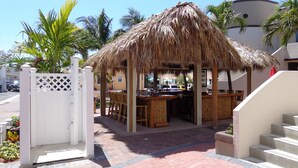 A thatched-roof outdoor bar area with a white fence and palm trees in the background.
