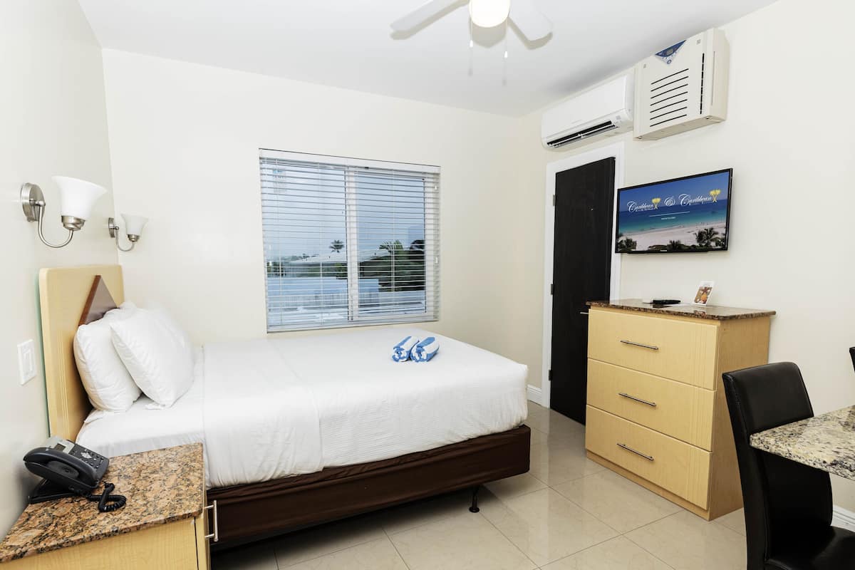 A hotel room with a bed, a desk with a telephone, a chair, a dresser, a window with blinds, an air conditioning unit, and a framed picture on the wall.