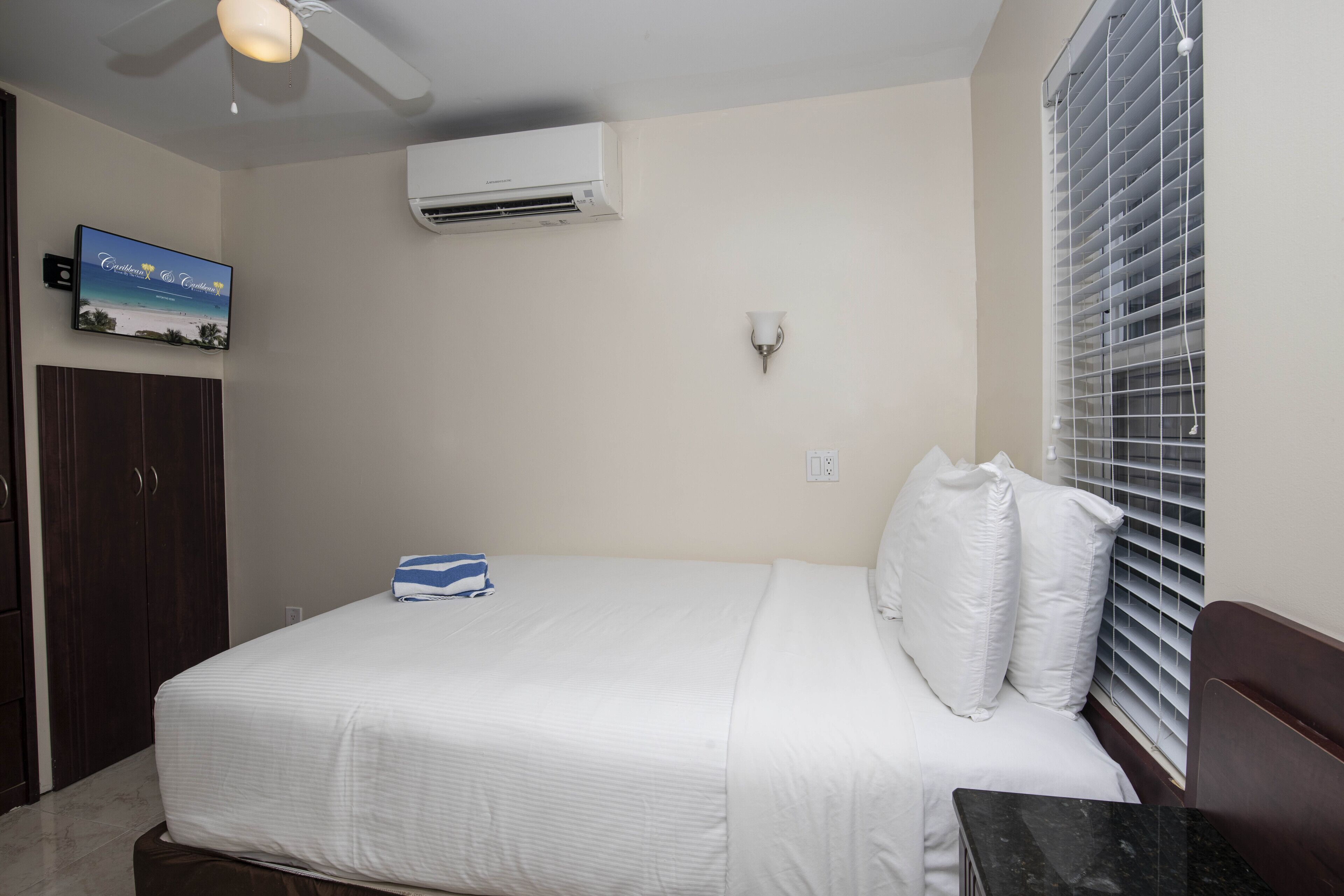 A bedroom with a bed, a ceiling fan, an air conditioning unit, a wall-mounted TV, and a window with blinds.