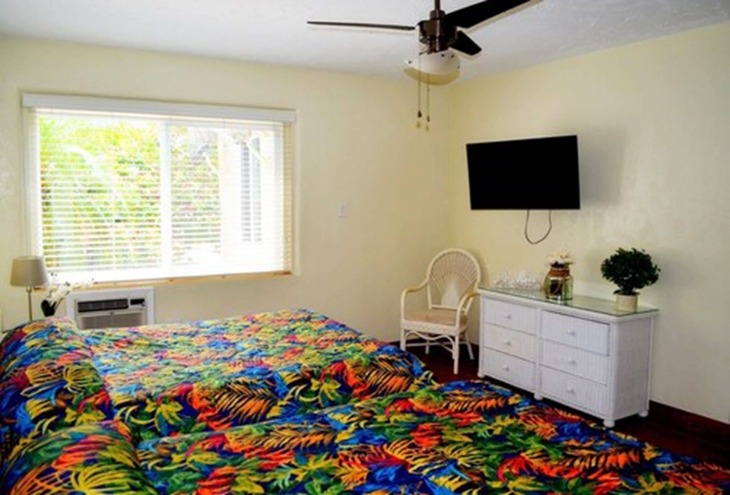 A room with a bed, a ceiling fan, a TV mounted on the wall, a window with blinds, a chair, and a dresser.