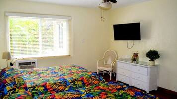 A room with a bed, a ceiling fan, a TV mounted on the wall, a window with blinds, a chair, and a dresser.