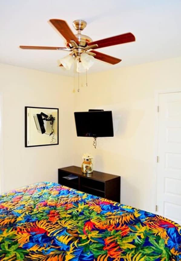 A bedroom with a ceiling fan, a flat-screen TV mounted on the wall, a bed with a colorful floral bedspread, and a small wooden nightstand with a lamp.