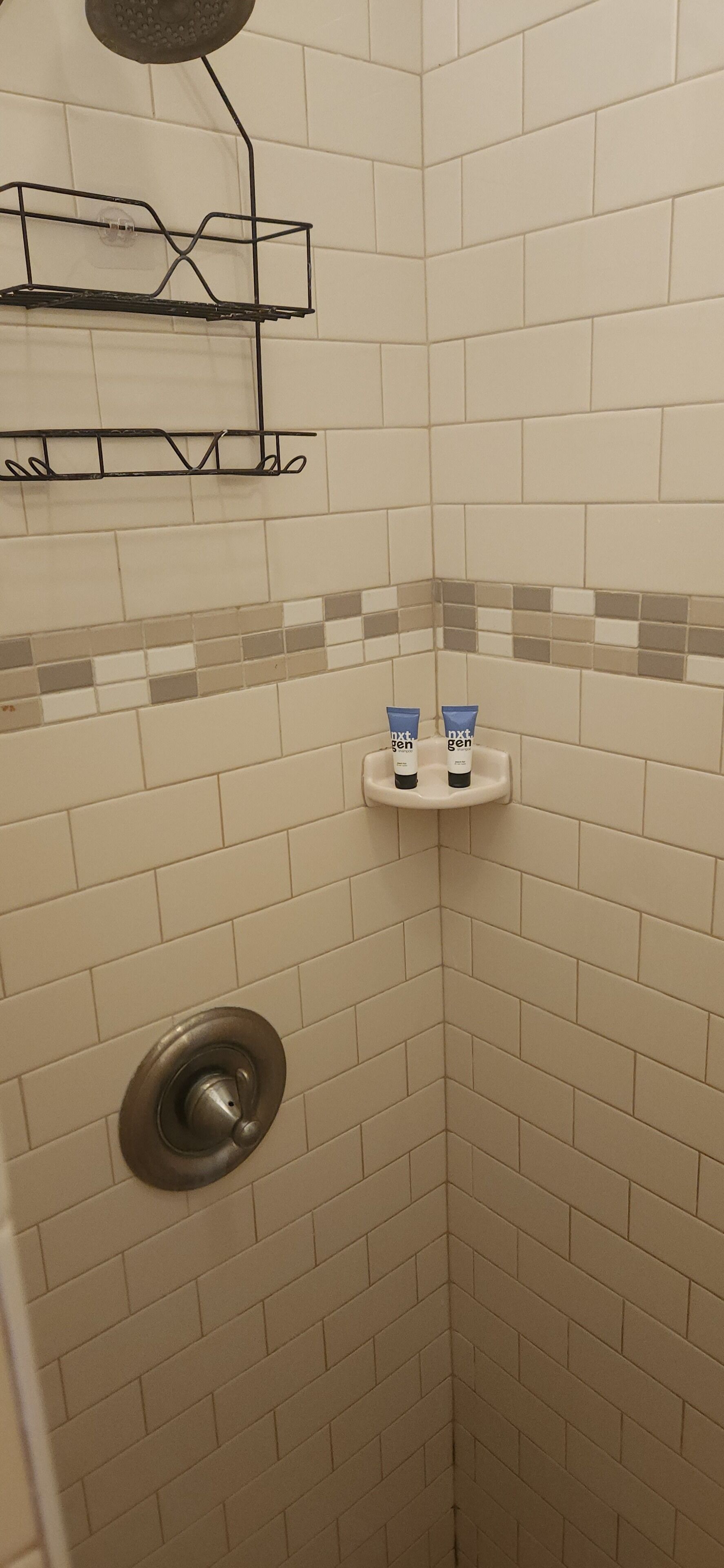 A shower area with beige tiles, a metal showerhead, and two blue toiletries on a white shelf.