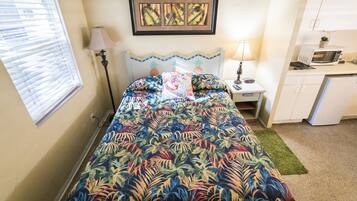 A bedroom with a bed, a nightstand, a lamp, a framed picture, a window with blinds, and a kitchenette area.
