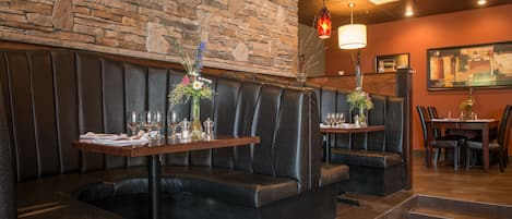 A restaurant with dark leather booths, stone walls, and hanging pendant lights. Tables are set with wine glasses and floral centerpieces.