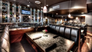 A bar area with a long counter, multiple bottles, and a television screen. There are leather booths and a table with a vase of flowers. - Podollan Inn & Spa Grande Prairie (Grande Prairie)