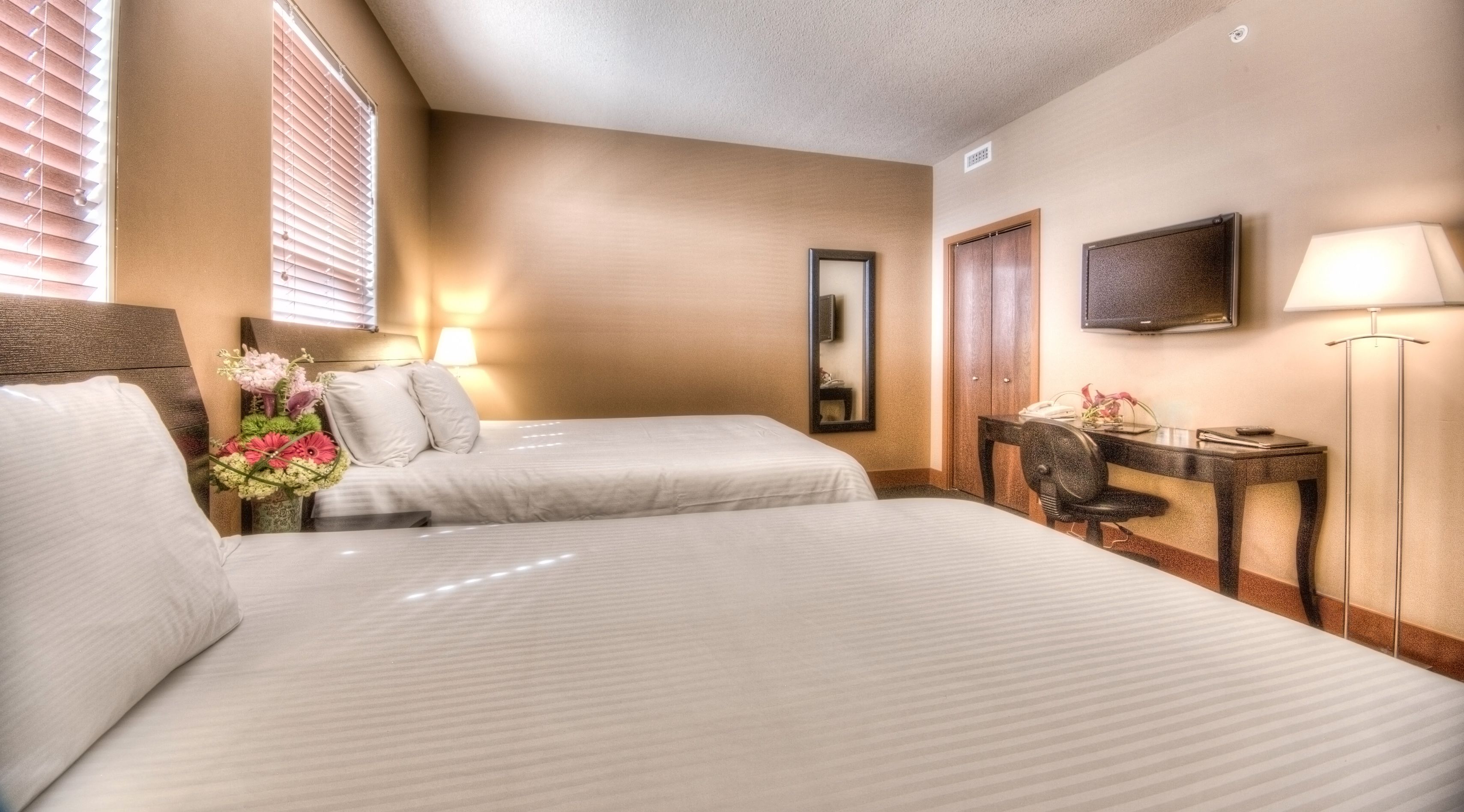 A hotel room with two beds, a desk, a chair, a lamp, a TV, and a mirror.