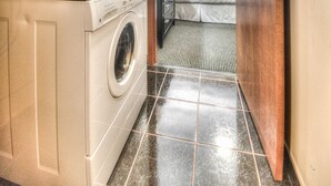 A laundry area with stacked washers and dryers, a bed with a striped bedspread, and a door leading to a bedroom. - Grand Inn & Residence (Grande Prairie)