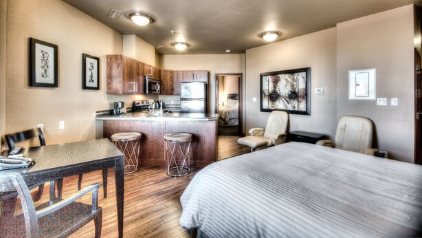 A hotel room with a bed, a desk with a chair, a kitchenette, and a bathroom visible through an open door. - Grand Inn & Residence (Grande Prairie)