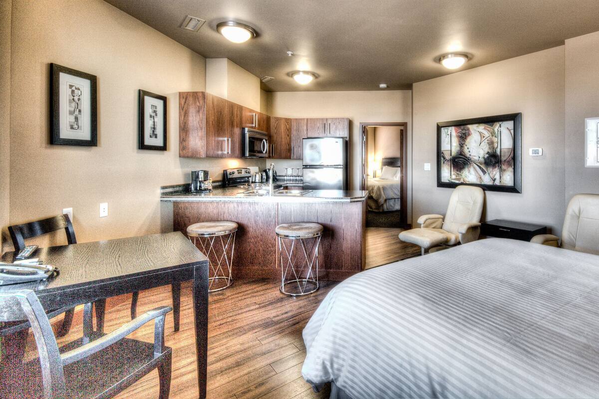A hotel room with a bed, a desk with a chair, a kitchenette, and a bathroom visible through an open door.
