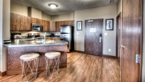 A kitchen with wooden cabinets, a stainless steel refrigerator, a microwave, a stove, and two bar stools. - Grand Inn & Residence (Grande Prairie)