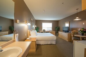 A hotel room with a large bed, a desk, a TV, a bathroom with a sink and mirror, and a window with blinds. - Prime Hotel (Fort McMurray)