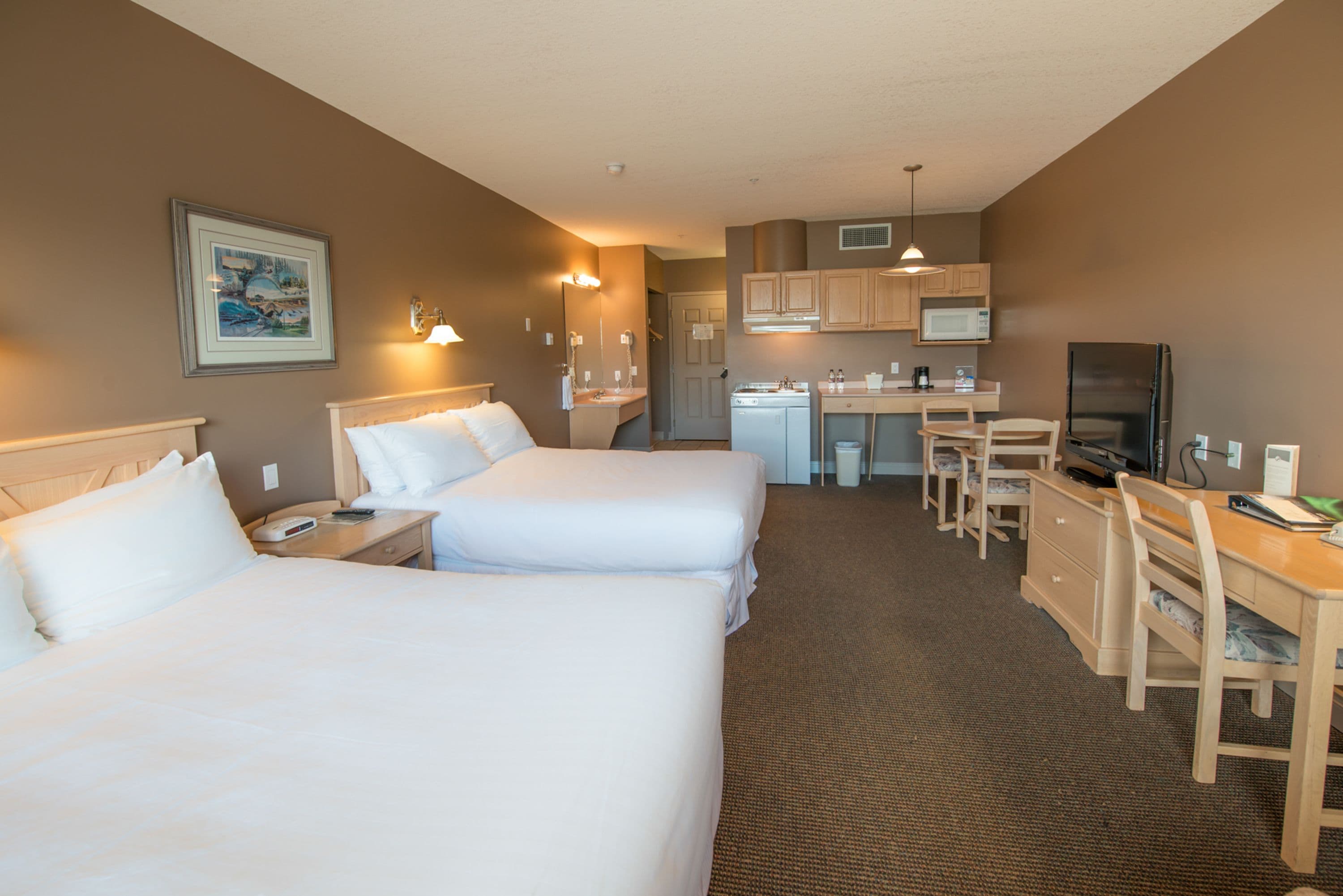 A hotel room with two beds, a desk, a chair, a television, and a kitchenette.