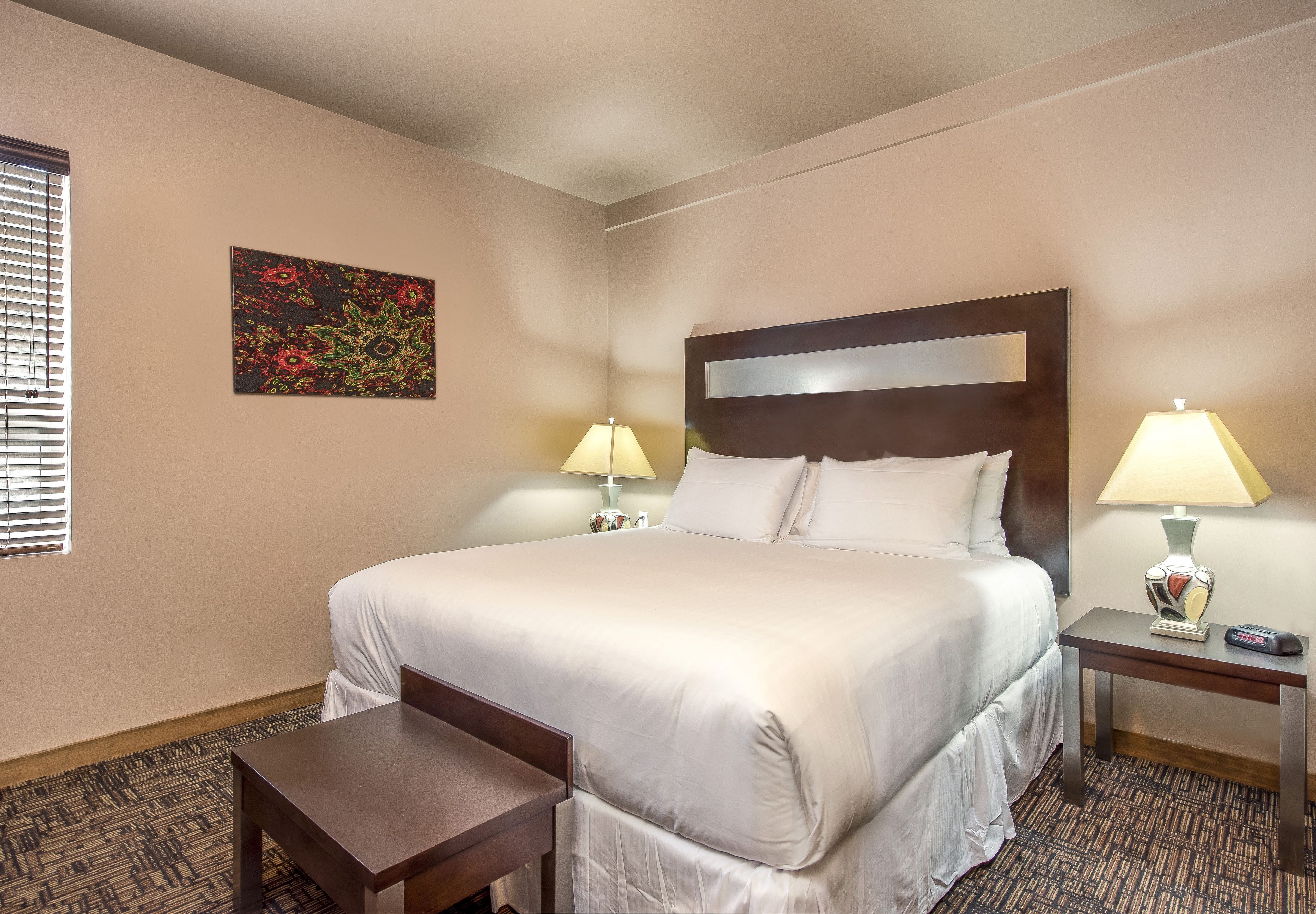 A hotel room with a large bed, two bedside tables with lamps, a small bench, and a decorative wall piece.