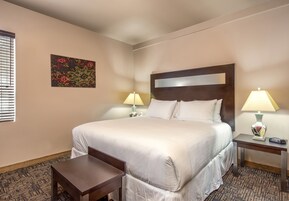 Una habitación de hotel con una cama grande, dos mesitas de noche con lámparas, un pequeño banco y un elemento decorativo en la pared.
