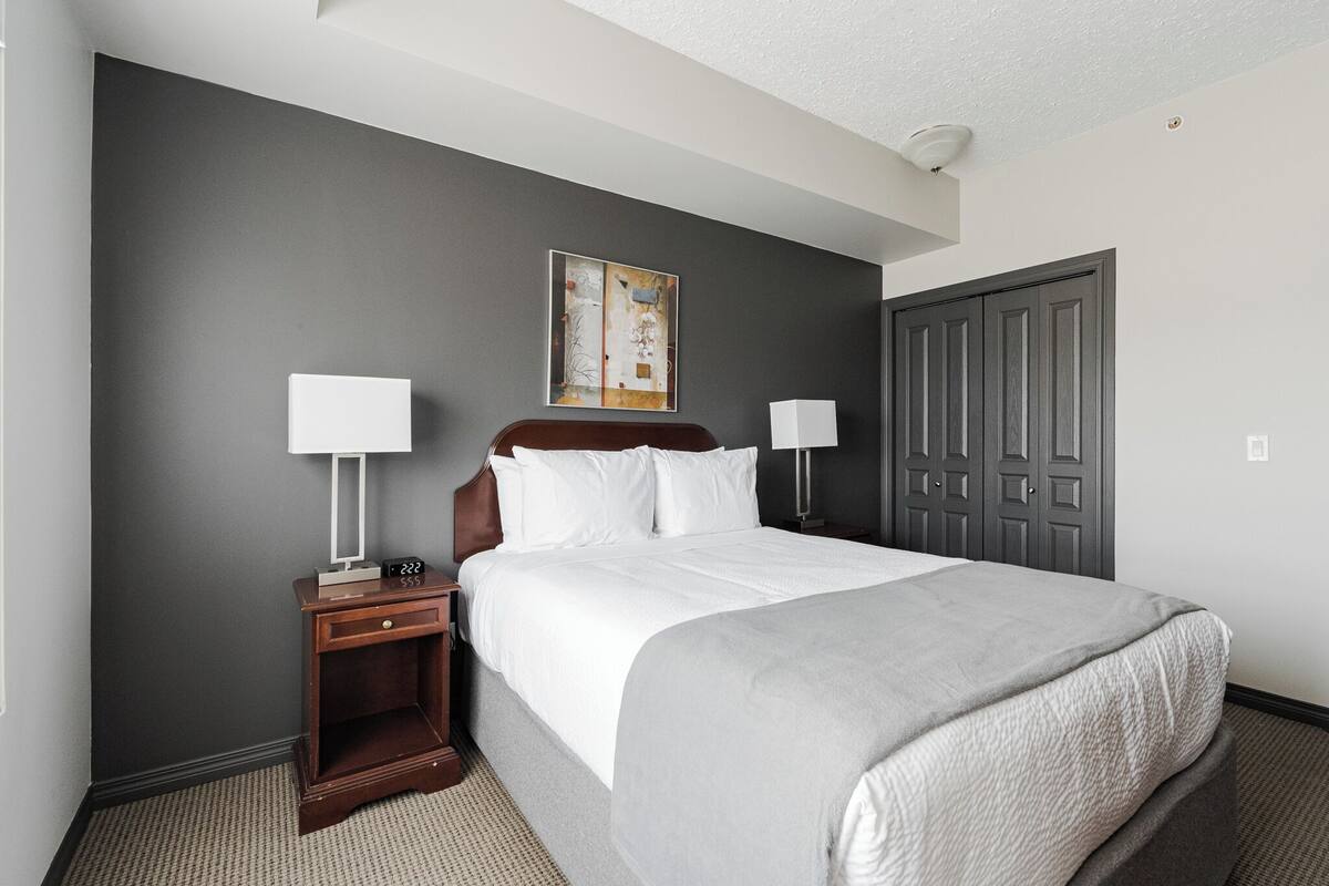 A hotel room with a large bed, two bedside tables with lamps, a gray wall, a door, and a painting above the bed.