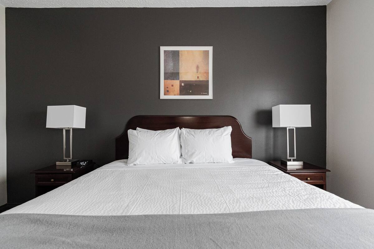 A hotel room with a large bed, two bedside tables with lamps, a nightstand with a clock, and a framed picture on the wall.