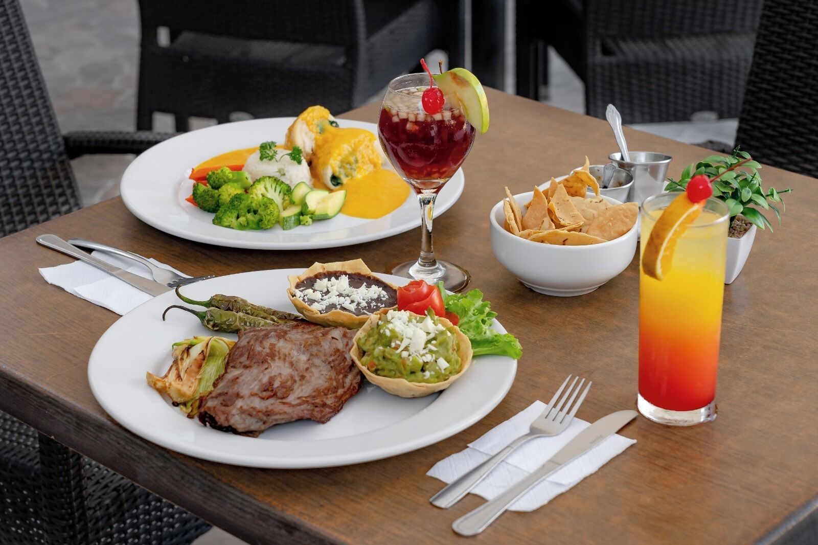 Ein Tisch mit Speisen, darunter gegrilltes Fleisch, Gemüse und ein Salat, sowie zwei Getränke und eine Schüssel Chips.