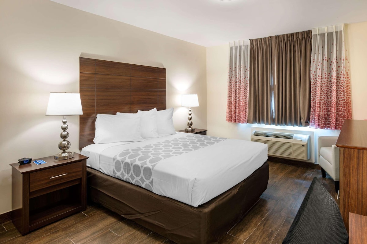 A hotel room with a large bed, two bedside tables with lamps, a window with curtains, and a built-in air conditioning unit.