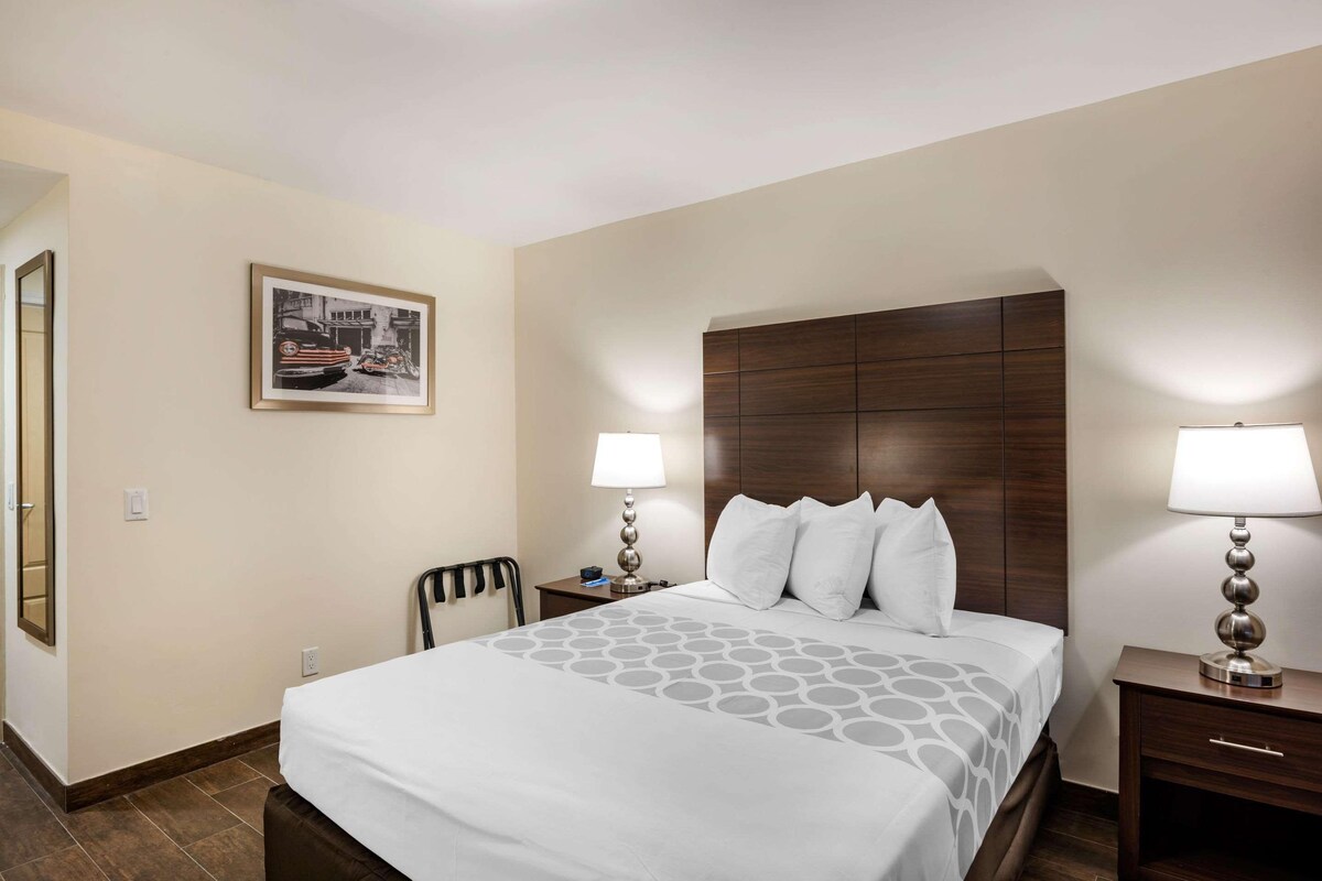A hotel room with a large bed, two bedside tables with lamps, a chair, a framed picture on the wall, and a wooden headboard.