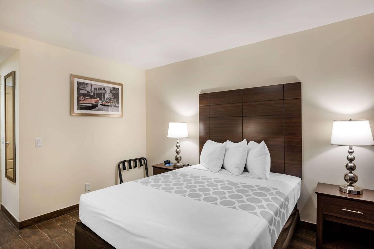 A hotel room with a large bed, two bedside tables with lamps, a chair, a framed picture on the wall, and a wooden headboard.