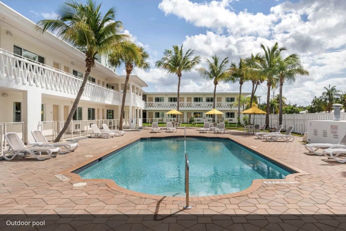 An outdoor pool with lounge chairs and umbrellas, surrounded by palm trees and a building with balconies.