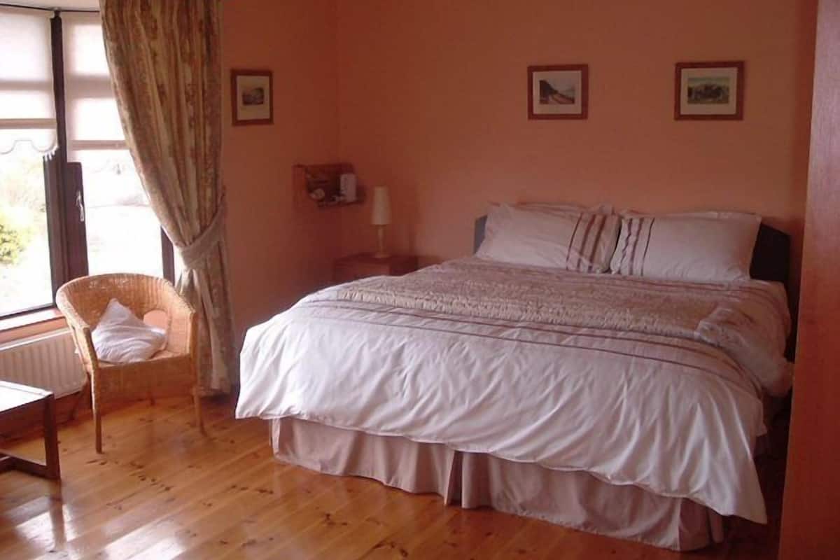 A bedroom with a bed, a chair, a window with curtains, and a wall with framed pictures.