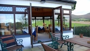 A covered outdoor seating area with wooden benches and tables, a view of a dining setup inside, and a scenic landscape visible through large windows. - Mourneview B&B (Carlingford)