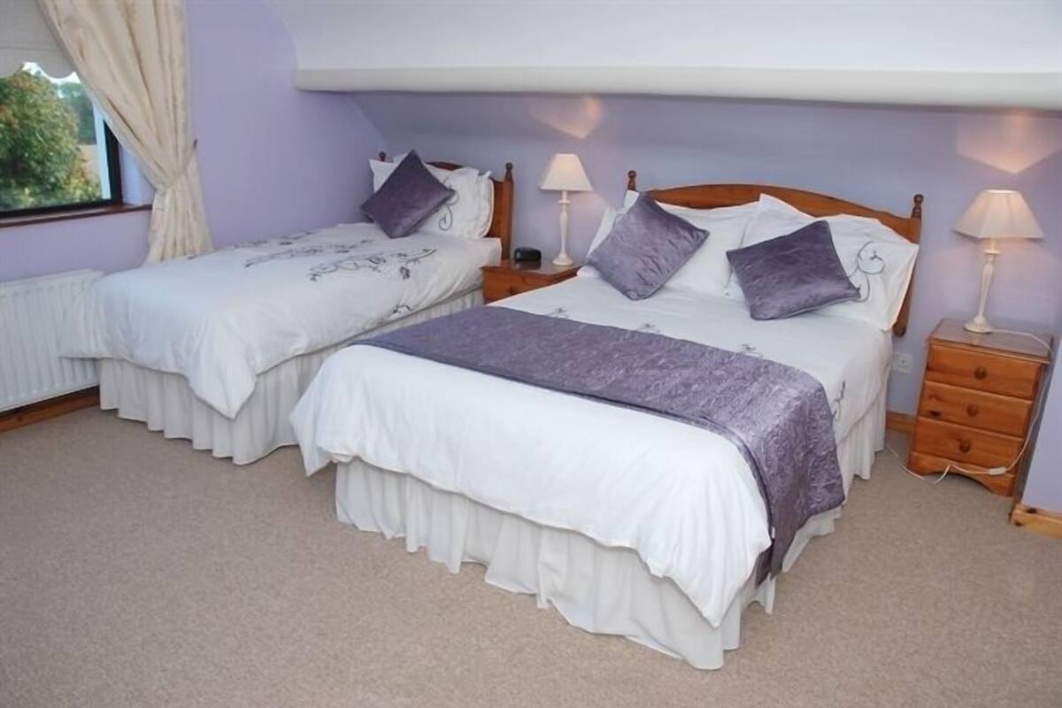 A bedroom with two single beds, each with a nightstand and lamp. The room has a window with curtains, a radiator, and a wooden headboard.