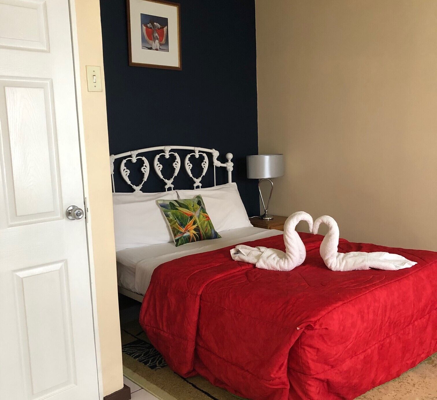 A hotel room with a bed, a red blanket, a white headboard, a pillow with a tropical design, a lamp, and a framed picture on the wall.