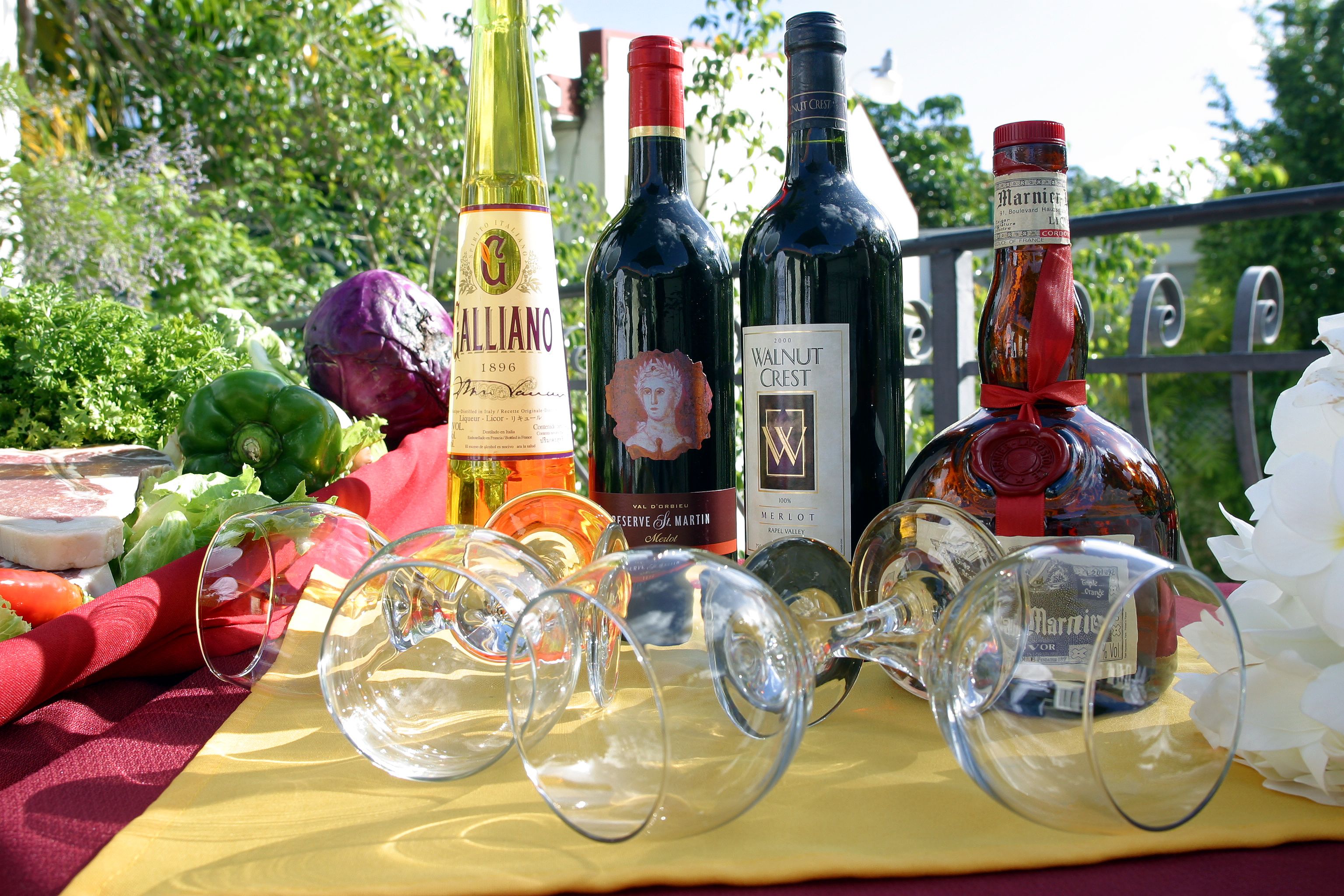 A table with wine bottles and glasses, a red cloth, and some vegetables.
