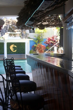 A bar area with a pool view, bar stools, and a decorative wall with a logo. - The Chancellor Hotel (Port of Spain)