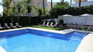 Piscina con sillas de descanso y sombrillas, rodeada de setos y palmeras.