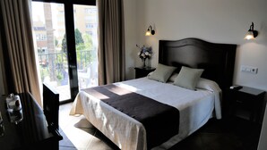 Un dormitorio con una cama grande, una mesita de noche con una lámpara, un florero con flores y vistas a un balcón.
