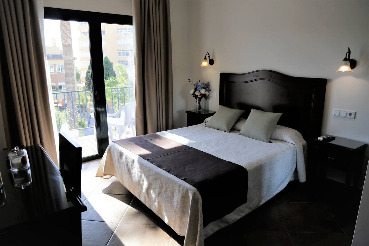 Un dormitorio con una cama grande, una mesita de noche con una lámpara, un florero con flores y vistas a un balcón.