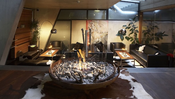 Un salon moderne doté d’un foyer central, de sièges confortables et offrant une vue sur le ciel grâce à une verrière.