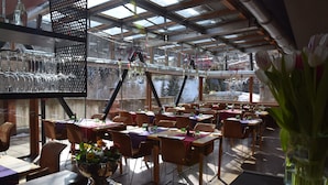 Un espace de restauration intérieur avec des tables et des chaises, un présentoir en verre contenant des verres à vin, et une vue sur l’extérieur.